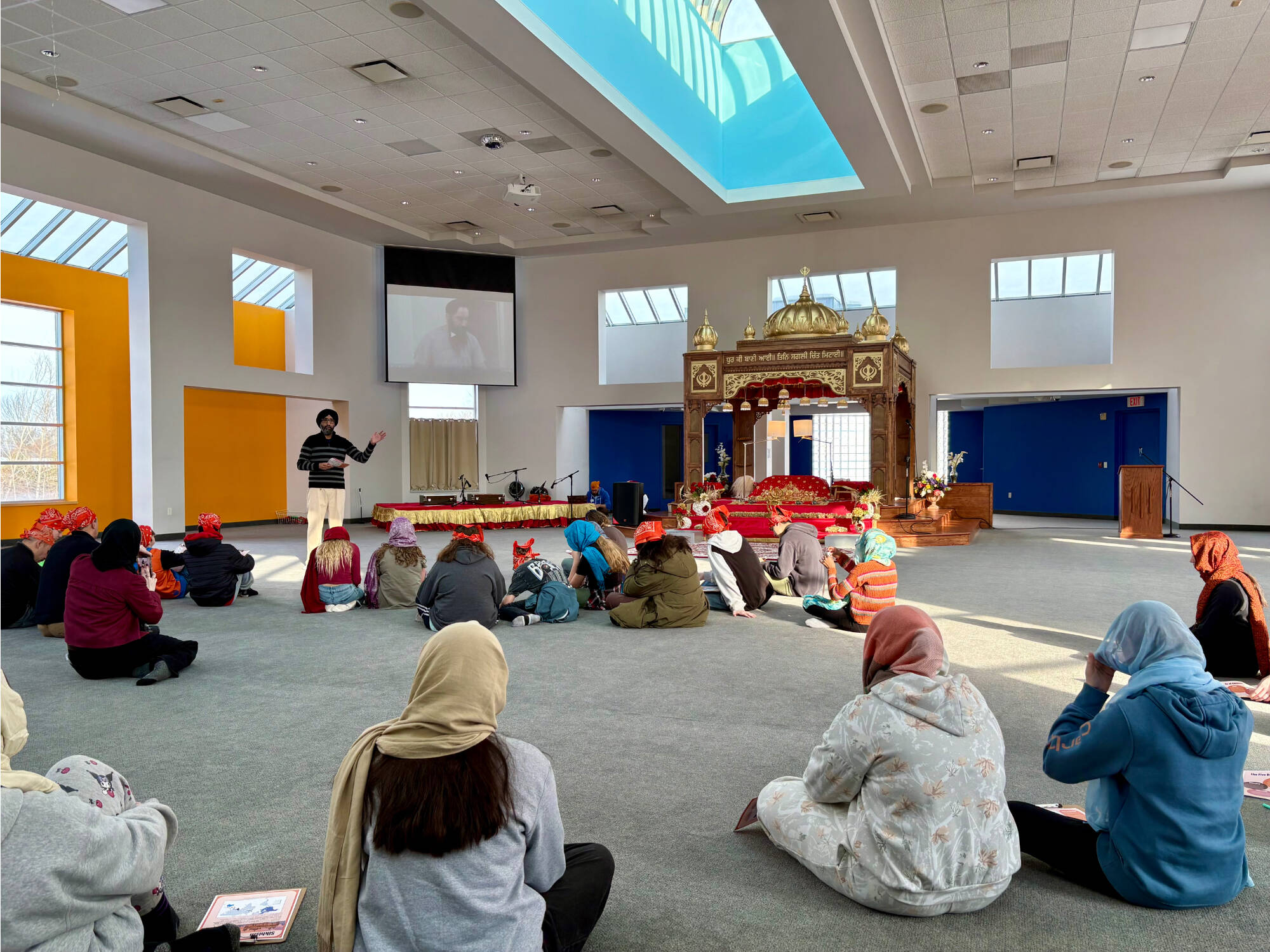 A wide shot of the gurdwara's primary worship all with students and a Sikh man gesturing